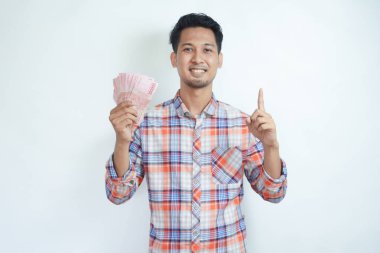 Adult Asian man smiling happy and pointing finger up while holding Indonesia paper money clipart