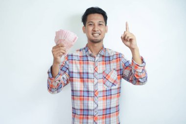 Adult Asian man smiling happy and pointing finger up while holding Indonesia paper money clipart