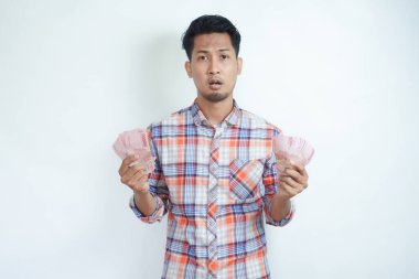 Adult Asian man holding money and looking camera with confused expression clipart