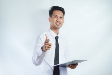 Genç Asyalı iş adamı elinde bir not defteri tutarken kamerayı işaret ediyor ve yüzünde bir gülümsemeyle ona bakıyordu. beyaz arkaplanda izole