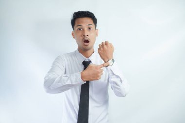 Yetişkin Asyalı bir adam sandalyede oturuyor ve kol saatini gösterirken kızgın yüz ifadesi sergiliyor.