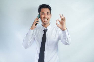 Adult Asian man smiling confident and give OK finger sign while holding a mobile phone clipart