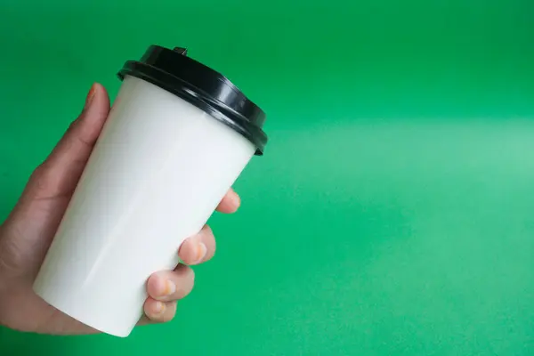 A hand holds an white disposable cup of tea or coffee on a green background. Blank space for product placement or advertising text.