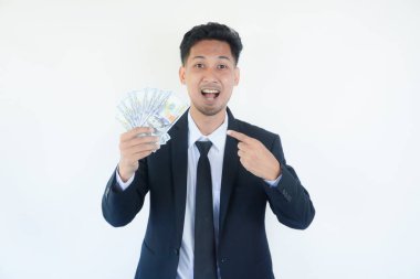 Takım elbiseli heyecanlı bir gencin portresi beyaz arka planda izole edilmiş bir sürü para banknotunu işaret ediyor.