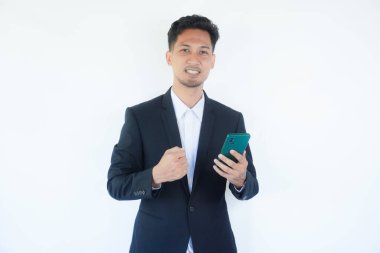 Adult Asian man wearing black suit and tie clenched fist while looking to his handphone clipart