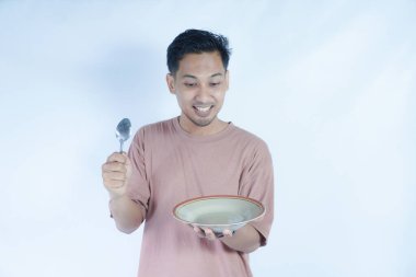 Pembe t-shirt giyen Endonezyalı bir adamın portresi, yeşil bir arka planda, bir kaşık ve tabakla mutlu poz verirken ve yemeğin tadını çıkarırken görülüyor..