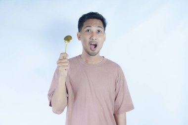 Tutkulu genç Asyalı adam beyaz arka planda izole edilmiş yemekleri tatmak için elinde pembe bir kaşık tutuyor. Halk Yaşam Tarzı Konsepti