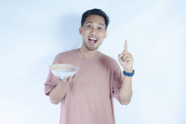 Pembe t-shirt giyen Endonezyalı bir adamın portresi. Beyaz bir arka planda, elinde tuttuğu boş bir kâseyi mutlulukla gösteriyor..