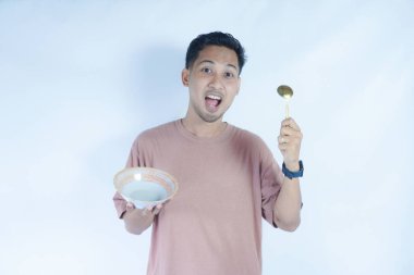 Pembe tişört giyen Endonezyalı bir adamın portresi, yeşil bir arka planda izole edilmiş bir kaşık ve kâseyle mutlu poz verirken ve yemeğin tadını çıkarırken görülüyor..