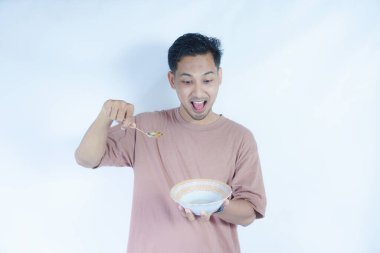 Pembe tişört giyen Endonezyalı bir adamın portresi, yeşil bir arka planda izole edilmiş bir kaşık ve kâseyle mutlu poz verirken ve yemeğin tadını çıkarırken görülüyor..