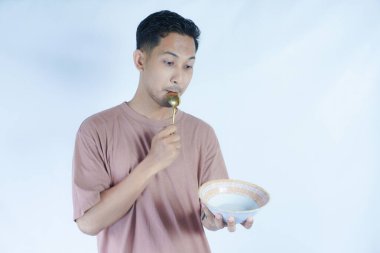 Pembe tişört giyen Endonezyalı bir adamın portresi, yeşil bir arka planda izole edilmiş bir kaşık ve kâseyle mutlu poz verirken ve yemeğin tadını çıkarırken görülüyor..