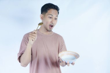 Pembe tişört giyen Endonezyalı bir adamın portresi, yeşil bir arka planda izole edilmiş bir kaşık ve kâseyle mutlu poz verirken ve yemeğin tadını çıkarırken görülüyor..