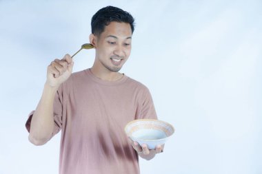 Pembe tişört giyen Endonezyalı bir adamın portresi, yeşil bir arka planda izole edilmiş bir kaşık ve kâseyle mutlu poz verirken ve yemeğin tadını çıkarırken görülüyor..