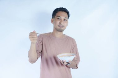 Pembe tişört giyen Endonezyalı bir adamın portresi, yeşil bir arka planda izole edilmiş bir kaşık ve kâseyle mutlu poz verirken ve yemeğin tadını çıkarırken görülüyor..