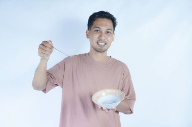 Pembe tişört giyen Endonezyalı bir adamın portresi, yeşil bir arka planda izole edilmiş bir kaşık ve kâseyle mutlu poz verirken ve yemeğin tadını çıkarırken görülüyor..