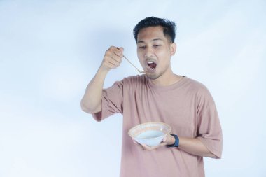Pembe tişört giyen Endonezyalı bir adamın portresi, yeşil bir arka planda izole edilmiş bir kaşık ve kâseyle mutlu poz verirken ve yemeğin tadını çıkarırken görülüyor..