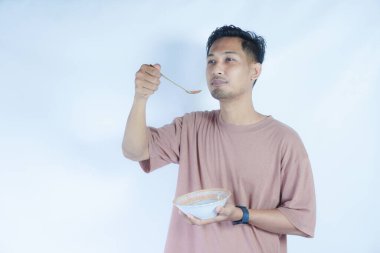 Pembe tişört giyen Endonezyalı bir adamın portresi, yeşil bir arka planda izole edilmiş bir kaşık ve kâseyle mutlu poz verirken ve yemeğin tadını çıkarırken görülüyor..