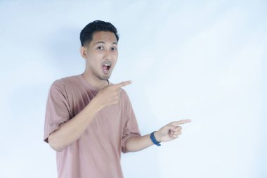 Pembe tişörtlü bir adam heyecanlı bir şekilde iki eliyle, ağzı açık bir şekilde şaşkınlıkla ve şaşkınlıkla onu işaret eder. Açık mavi arka plan, onun hareketli ve coşkulu ifadesini vurguluyor..
