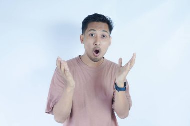 Pembe tişörtlü Asyalı bir adam, düz bir arkaplan karşısında duruyor, şaşırmış bir ifade ve yüzüne yakın duran eller. Görüntü bir şok ya da şaşkınlık anını yakalar..