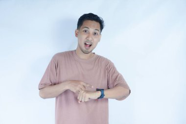 Pembe tişörtlü Asyalı bir adam endişeli ve sabırsız bir ifadeyle saatine bakar. Zaman konusunda endişeli olduğunu gösterir. Açık mavi arka plan odaklandığı şeyi vurguluyor.