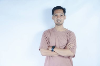 Pembe gömlekli Asyalı bir adam kollarını kavuşturup kameraya samimi bir şekilde gülümsüyor. Açık mavi arka plan onun rahat ve cana yakın tavrını vurguluyor..