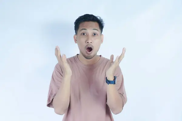 stock image Asian man in a pink t-shirt, standing against a plain background, with an expression of surprise and hands raised near his face. The image captures a moment of shock or astonishment.