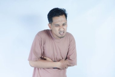 Pembe gömlekli bir adam, midesi ağrıyan bir yüz ifadesiyle karnını tutar. Bu da mide ağrısı ya da rahatsızlık gösterir. Açık mavi arka plan sıkıntısını vurguluyor.