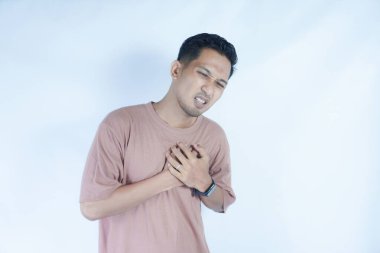 Pembe gömlekli bir adam göğsünü iki eliyle kavrıyor. Acı dolu bir yüz ifadesi sergiliyor. Muhtemelen göğüs ağrısı ya da rahatsızlığı gösteriyor. Açık mavi arka plan sıkıntısını vurguluyor.