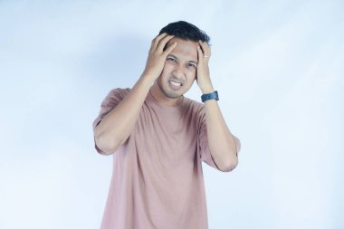 Pembe gömlekli bir adam başını acı dolu bir ifadeyle tutar. Baş ağrısı ya da rahatsızlık gösterir. Açık mavi arka plan sıkıntısını vurguluyor ve duruşunu ve ifadesini vurguluyor.