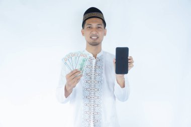 Asyalı Müslüman adam elinde para tutarken gülümsüyor ve boş bir cep telefonu ekranı gösteriyor.