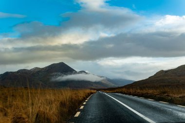 Bulutlu bir gökyüzünün altındaki sisli dağlara doğru uzanan boş bir yol, her iki tarafında çimen tarlaları olan..