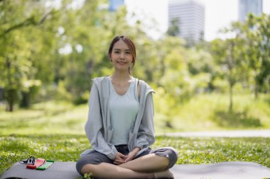 Genç yetişkin Asyalı kadın, parktaki yoga ve meditasyon uygulamalarıyla yeniden canlandırmayı keşfeder. Fiziksel ve zihinsel yeteneklerin hizalanması enerji seviyelerini, genel canlılığı ve refahı onarması