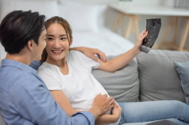 Asyalı çiftler ultrason görüntülerini tararken hamileliğin coşkusunu ve heyecanını kucaklarlar. Bebeğin rahmindeki sağlık, cinsiyet ve gelişimi ortaya çıkar. Mutluluk ve memnuniyet aile içinde bağı güçlendirir.