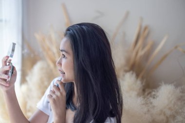 Diş telleri olan genç bir Asyalı kız makyaj yapıyor, rahat, aydınlık bir odada el kamerasına bakıyor..