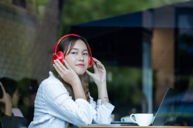 Düşünceli bir Asyalı kadın, rahat bir kafede laptopuyla çalışırken kırmızı kulaklıkla müzik dinliyor..