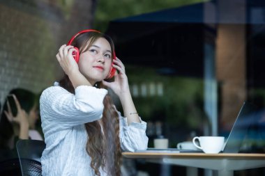 Düşünceli bir Asyalı kadın, rahat bir kafede laptopuyla çalışırken kırmızı kulaklıkla müzik dinliyor..