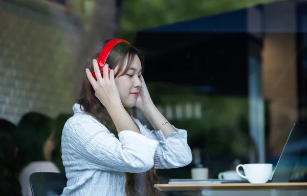 A contemplative Asian woman listens to music on red headphones while working on her laptop in a cozy cafe setting.
