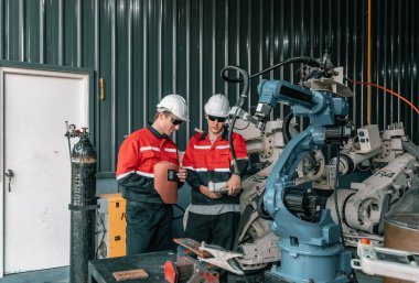 İki robotik kaynak teknisyeni, bir atölyede otomatik kaynak görevi için endüstriyel bir robotu programlıyor..