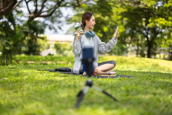 Bir kadın sosyal medya etkeni sohbetin ortasında, yemyeşil bir çimenlikte otururken çevrimiçi izleyicilerine protein tozu içeceği sergilerken yakalanıyor..