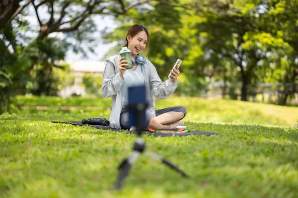 Bir kadın sosyal medya etkeni sohbetin ortasında, yemyeşil bir çimenlikte otururken çevrimiçi izleyicilerine protein tozu içeceği sergilerken yakalanıyor..