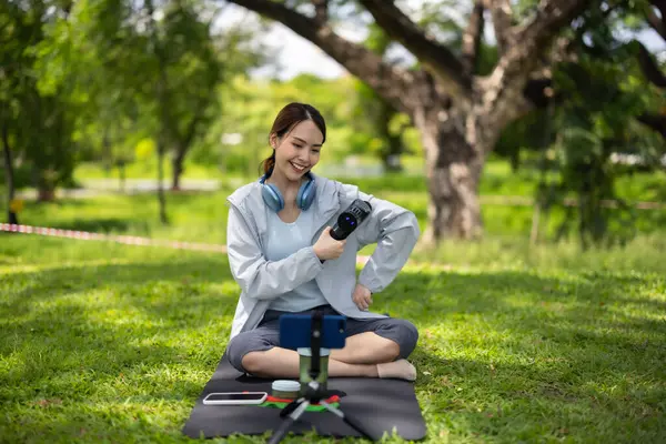 Bir kadın sosyal medya etkeni yemyeşil bir parkta bir mindere oturmuş, izleyicileri için gösteriyi kaydederken masaj tabancasının nasıl kullanılacağını gösteriyor..