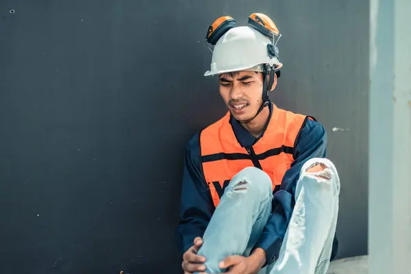 Asian HVAC engineer suffered serious leg injury on the job. Urgent first aid and assistance from coworkers is essential. Expressions of pain on face convey physical and emotional response.