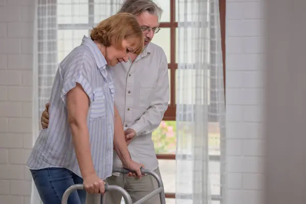 An elderly couple uses walking aids while assisting each other in an indoor setting, highlighting their mutual care.