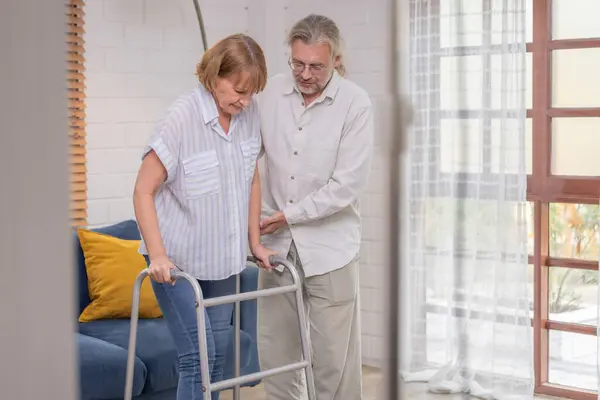 An elderly couple uses walking aids while assisting each other in an indoor setting, highlighting their mutual care.
