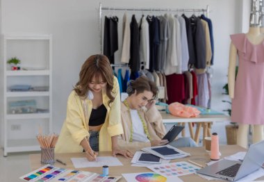 İki moda tasarımcısı, kumaş örnekleri, renk paletleri ve elbise raflarıyla çevrili yaratıcı çalışma alanlarının keyfini çıkarıyorlar. Bir tasarımcı kulaklıkla müzik dinlerken, diğeri tablete dalmış durumda..