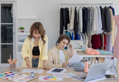 İki moda tasarımcısı, kumaş örnekleri, renk paletleri ve elbise raflarıyla çevrili yaratıcı çalışma alanlarının keyfini çıkarıyorlar. Bir tasarımcı kulaklıkla müzik dinlerken, diğeri tablete dalmış durumda..