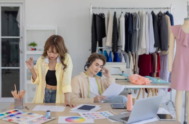 İki moda tasarımcısı, kumaş örnekleri, renk paletleri ve elbise raflarıyla çevrili yaratıcı çalışma alanlarının keyfini çıkarıyorlar. Bir tasarımcı kulaklıkla müzik dinlerken, diğeri tablete dalmış durumda..