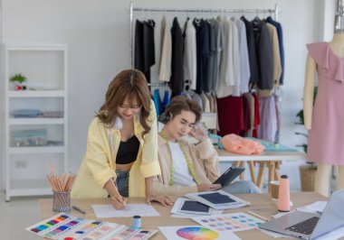 İki moda tasarımcısı, kumaş örnekleri, renk paletleri ve elbise raflarıyla çevrili yaratıcı çalışma alanlarının keyfini çıkarıyorlar. Bir tasarımcı kulaklıkla müzik dinlerken, diğeri tablete dalmış durumda..