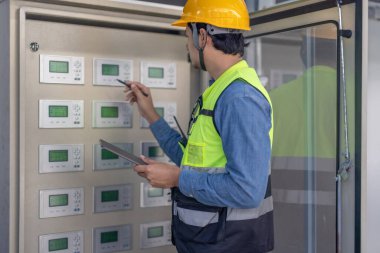 Sert şapka ve güvenlik yeleği giymiş bir havalandırma teknisyeni birden fazla ekranı olan kontrol panelini kontrol ediyor. Teknisyen bir kalem kullanıyor ve elinde bir tablet tutuyor. Düzgün işlevsellik ve sistem verimliliği sağlıyor.