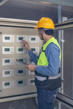 Sert şapka ve güvenlik yeleği giymiş bir havalandırma teknisyeni birden fazla ekranı olan kontrol panelini kontrol ediyor. Teknisyen bir kalem kullanıyor ve elinde bir tablet tutuyor. Düzgün işlevsellik ve sistem verimliliği sağlıyor.